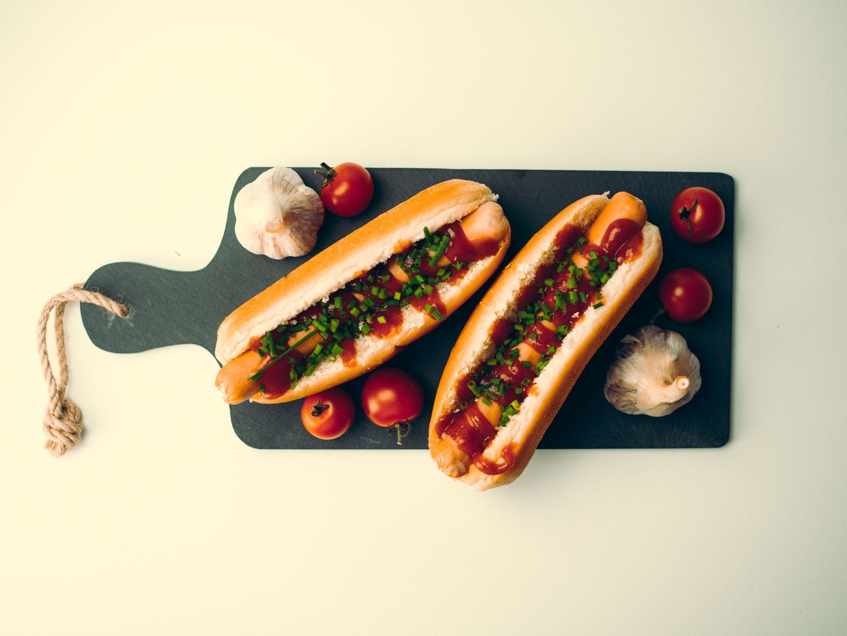 Two hotdogs with tomatoes and garlic on a dark chopping board